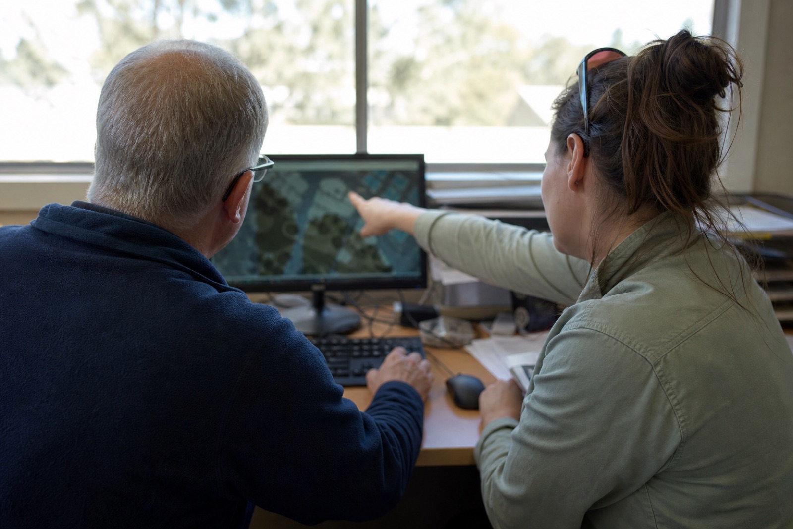 Two colleagues at one desk, the older one at the keyboard while a colleague points at something on the screen.