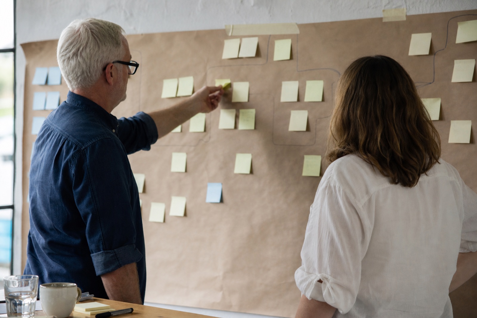 Two people at a wall of blank sticky notes and butcher's paper, mapping out a process by hand with their sleeves rolled up.