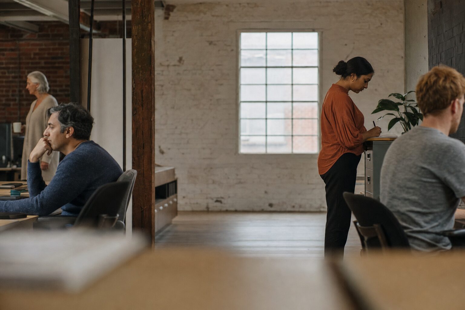 A converted warehouse office early in the morning. A man in a slate blue jumper sits sideways at a desk with his chin on his hand, a woman with silver hair stands further back holding a mug, a woman in a terracotta blouse writes on a pad resting on a filing cabinet, and a man in a grey t-shirt sits with his back to the room. The middle of the room is empty pale brick and one tall window.